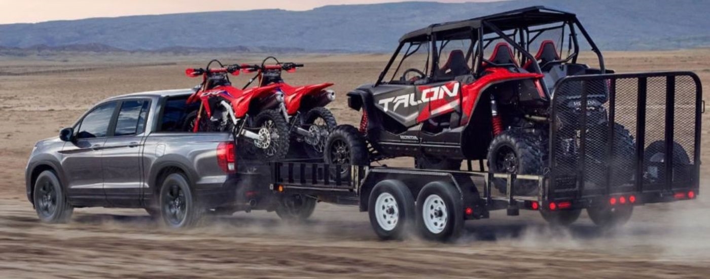 Side view of a gray 2026 Honda Ridgeline towing off-road recreational vehicles.