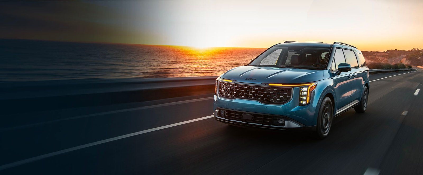 2027 Kia Carnival in blue driving on a coastal highway at sunset