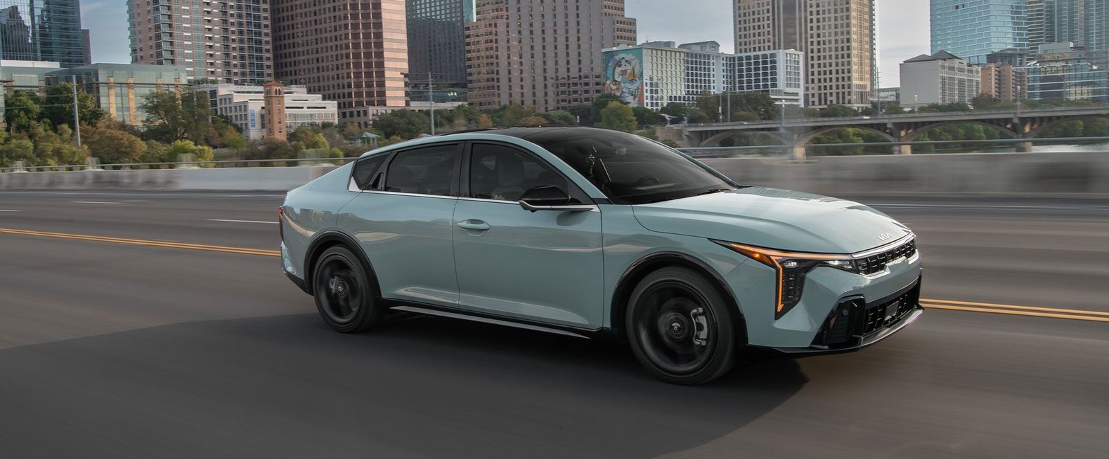 Kia K4 sedan in silver driving past a city skyline