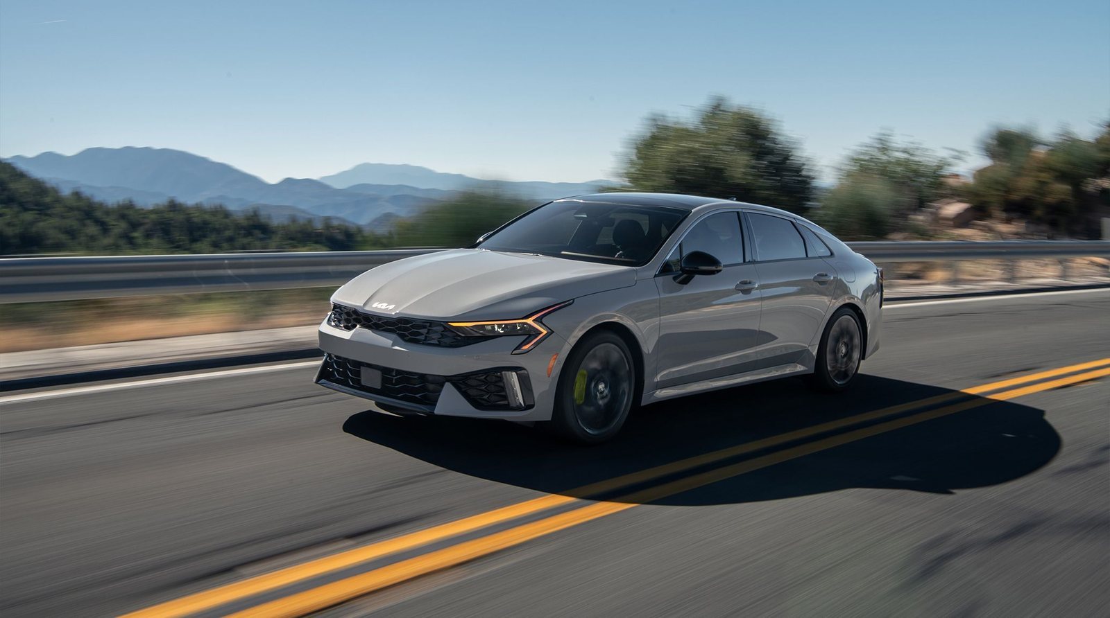 2027 Kia K5 in silver driving on a two-lane mountain highway
