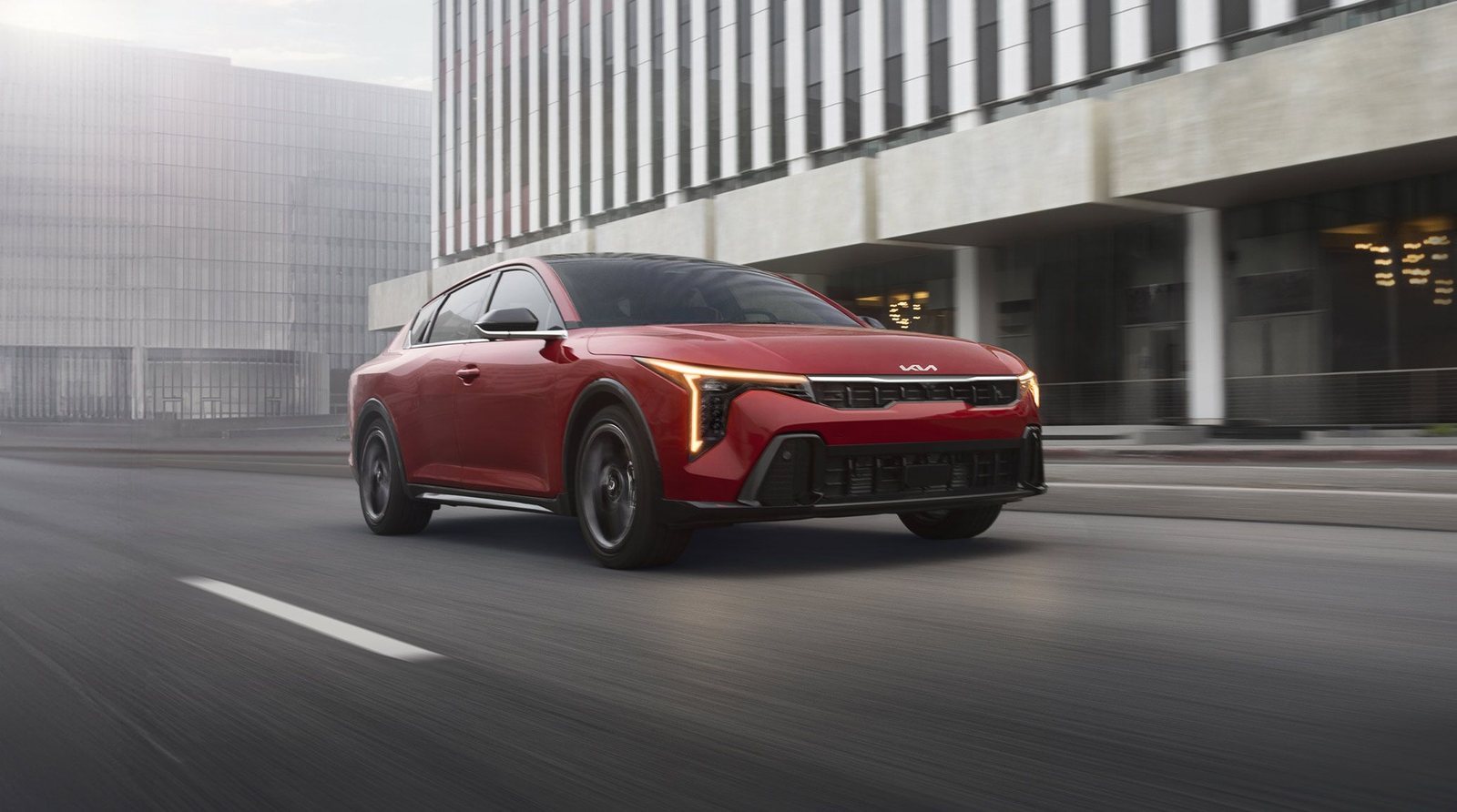 Kia K4 sedan in red driving past a modern office building