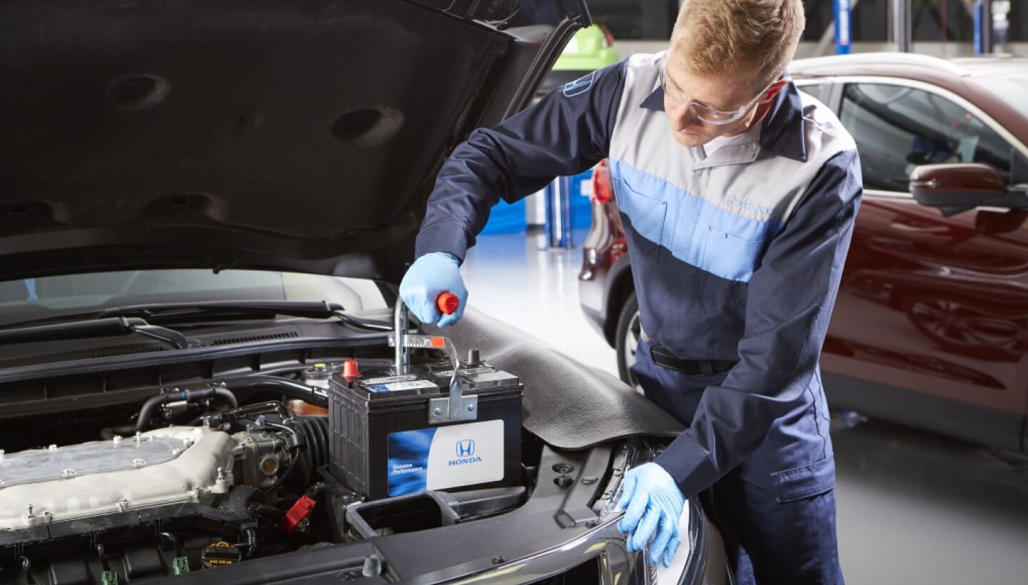A Honda Certified Technician installing a new battery under the open hood of a Honda vehicle.
