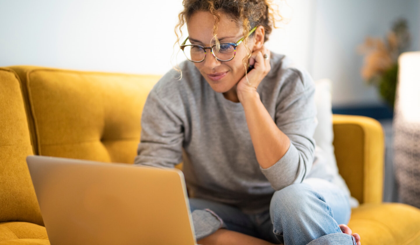A woman smiles while using her laptop on a yellow couch.