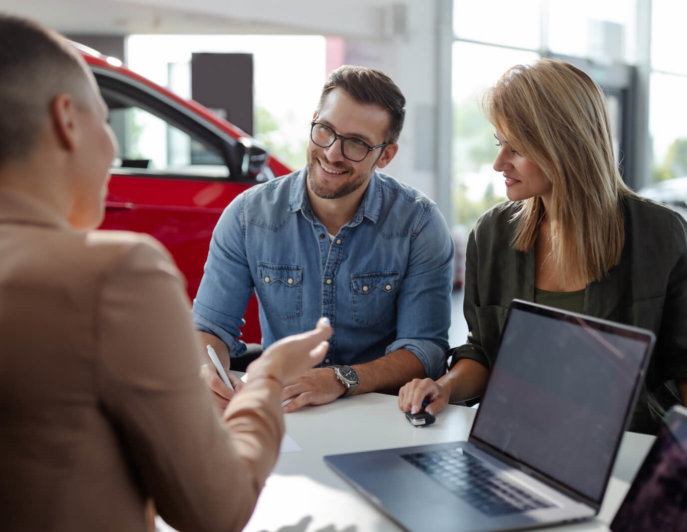 A sales representative assists a pair of customers at a desk in a sunlit dealership.