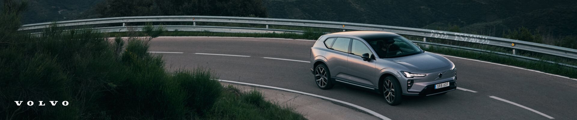 Front three-quarter view of a silver Volvo EX60 driving along a winding mountain road at sunset.   