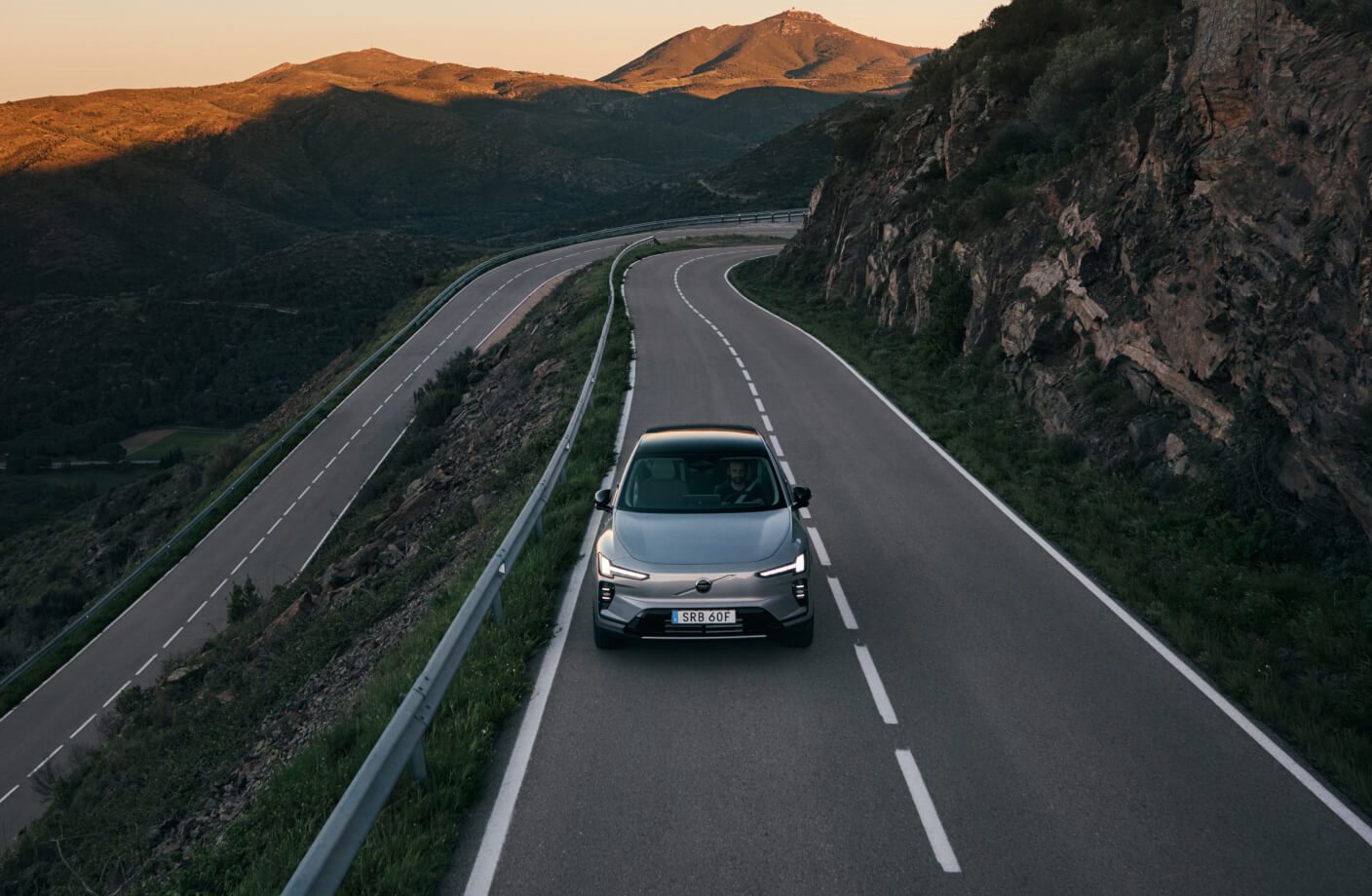 High-angle front view of a silver Volvo EX60 driving along a winding mountain road at sunset.