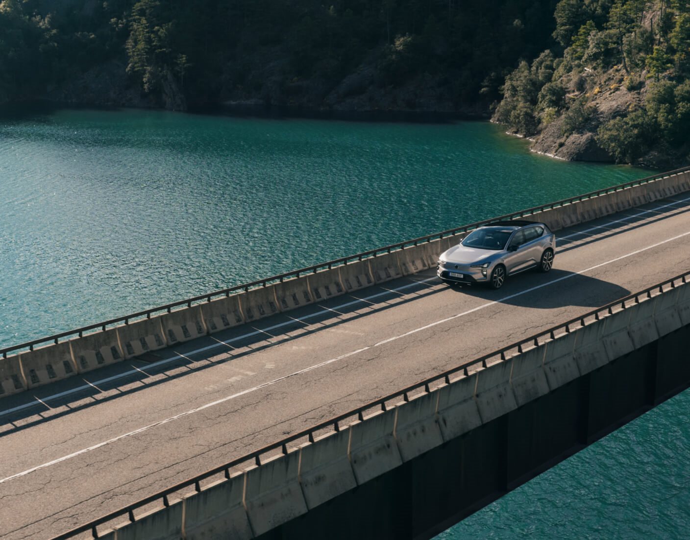 Aerial view of a silver Volvo EX60 crossing a bridge over a lake.   
L3: Rear side view of a dark green Volvo EX60 plugged into a charger beside a modern fluted wall.   