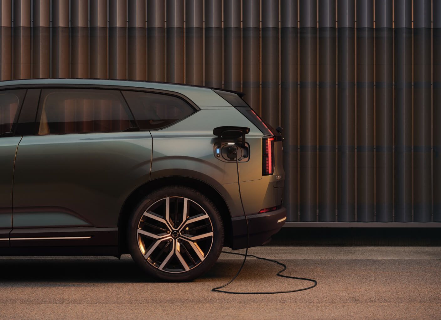 Rear side view of a dark green Volvo EX60 plugged into a charger beside a modern fluted wall.   
