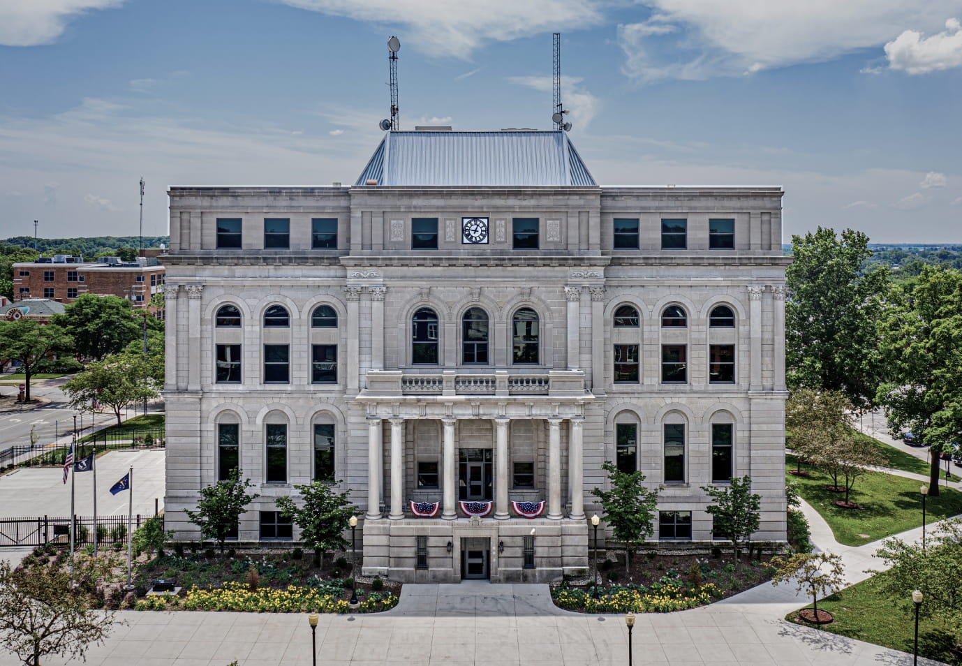 Front view of a historic stone courthouse featuring a central clock, arched windows, a columned entrance, patriotic bunting, and landscaped grounds.