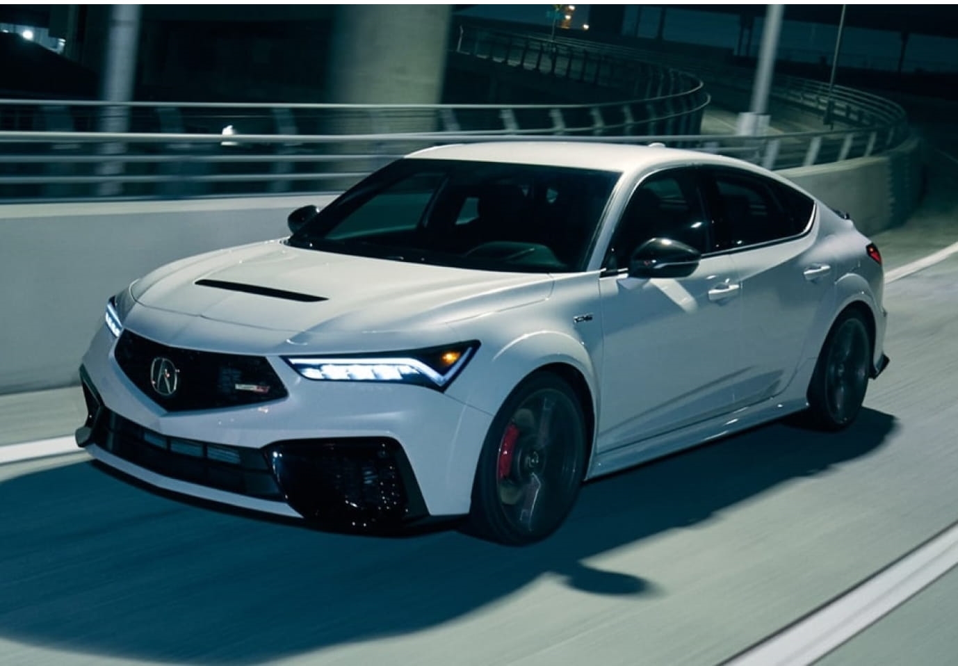 White Acura performance sedan driving along a curved urban roadway, featuring illuminated LED headlights, black alloy wheels, and sporty exterior styling.