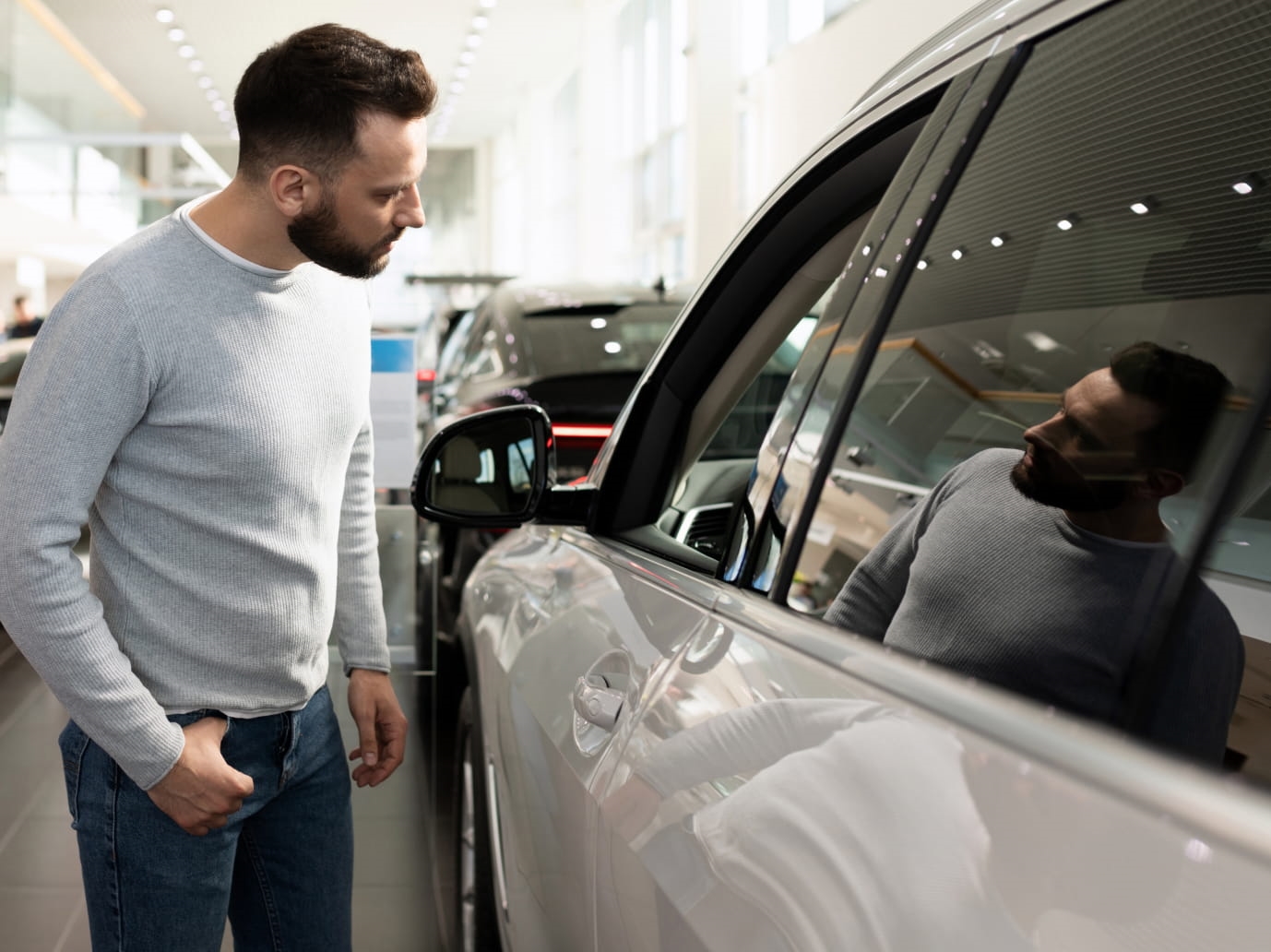 Man examining the side window of a white SUV inside a brightly lit car dealership showroom, with his reflection visible in the glass.