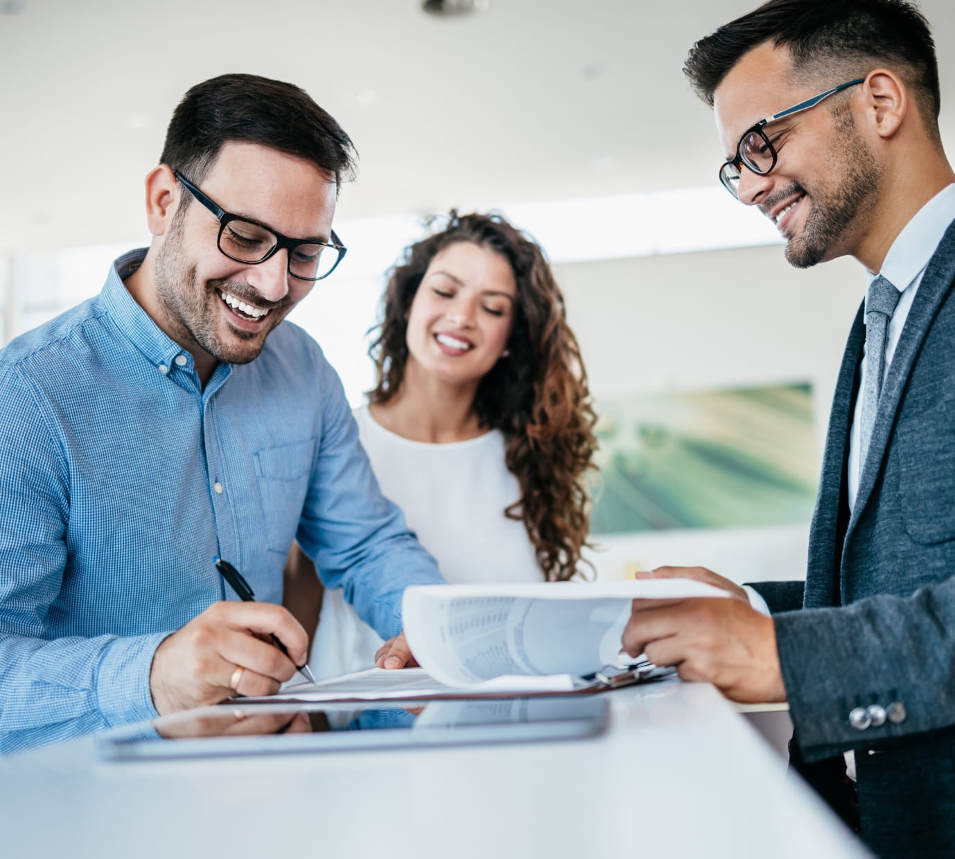 Smiling customer signing paperwork at a desk while a woman looks on and a sales representative assists with the documents.