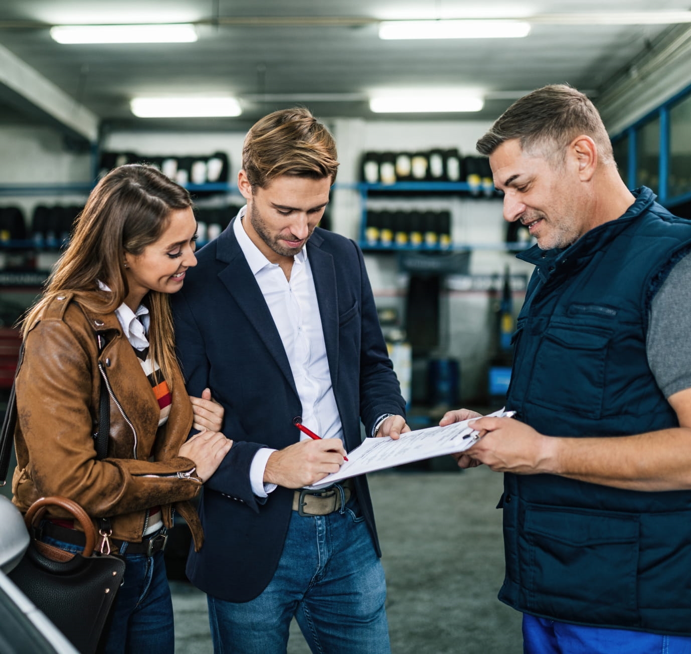 Customer signing paperwork on a clipboard while accompanied by a woman and assisted by a service representative inside an automotive service center.